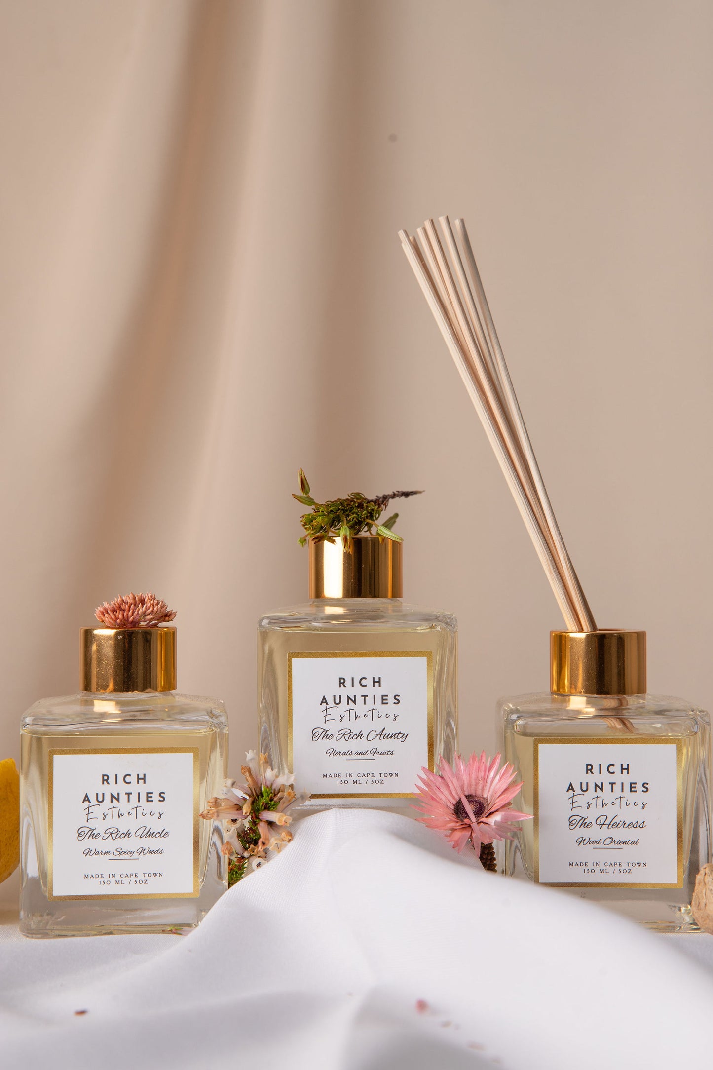 Reed diffusers in clear glass bottle and gold-colored cap, containing a diffuser fragrance and accompanied by several light beige reeds tied with flowers 