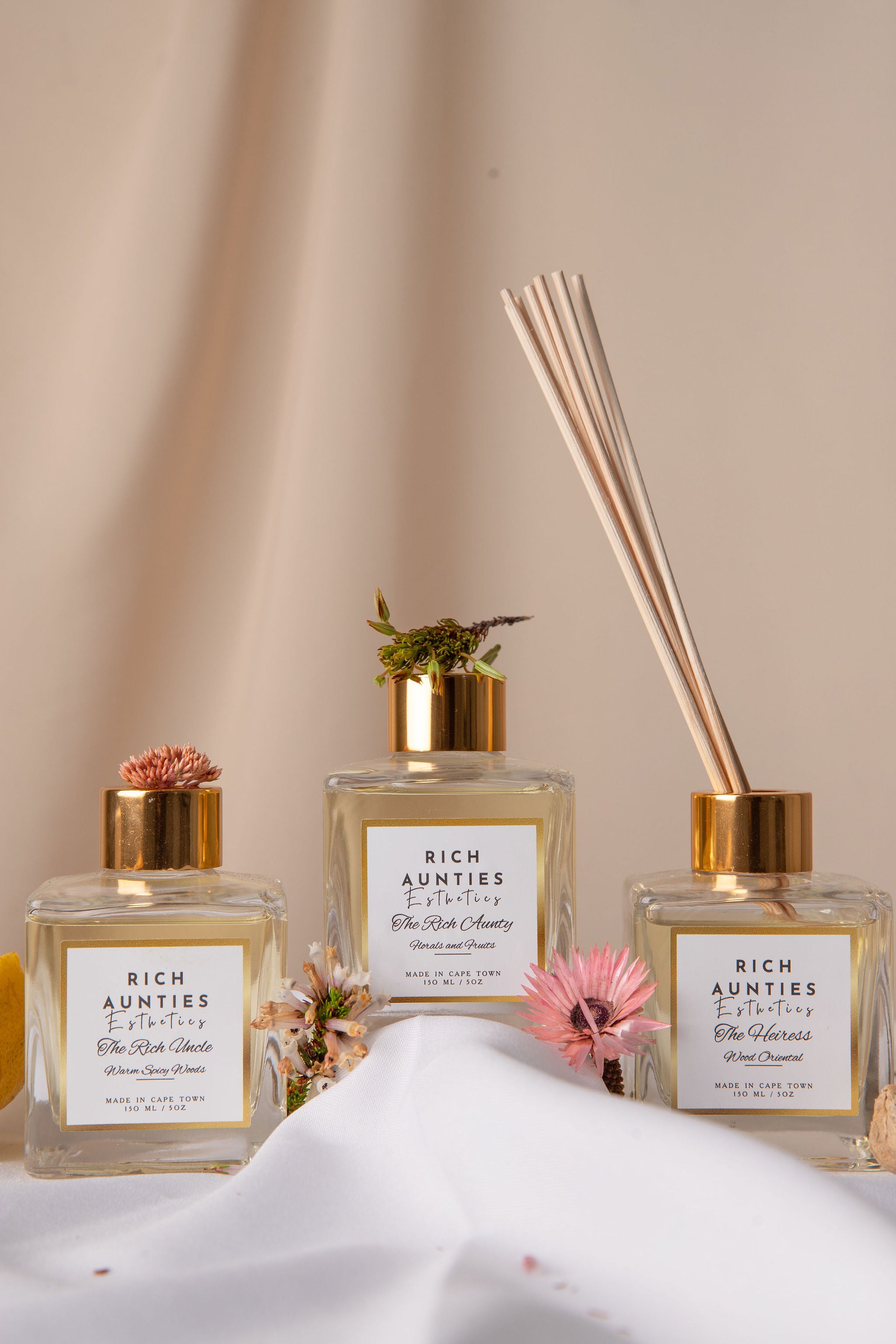 Reed diffusers in clear glass bottle and gold-colored cap, containing a diffuser fragrance and accompanied by several light beige reeds tied with flowers 