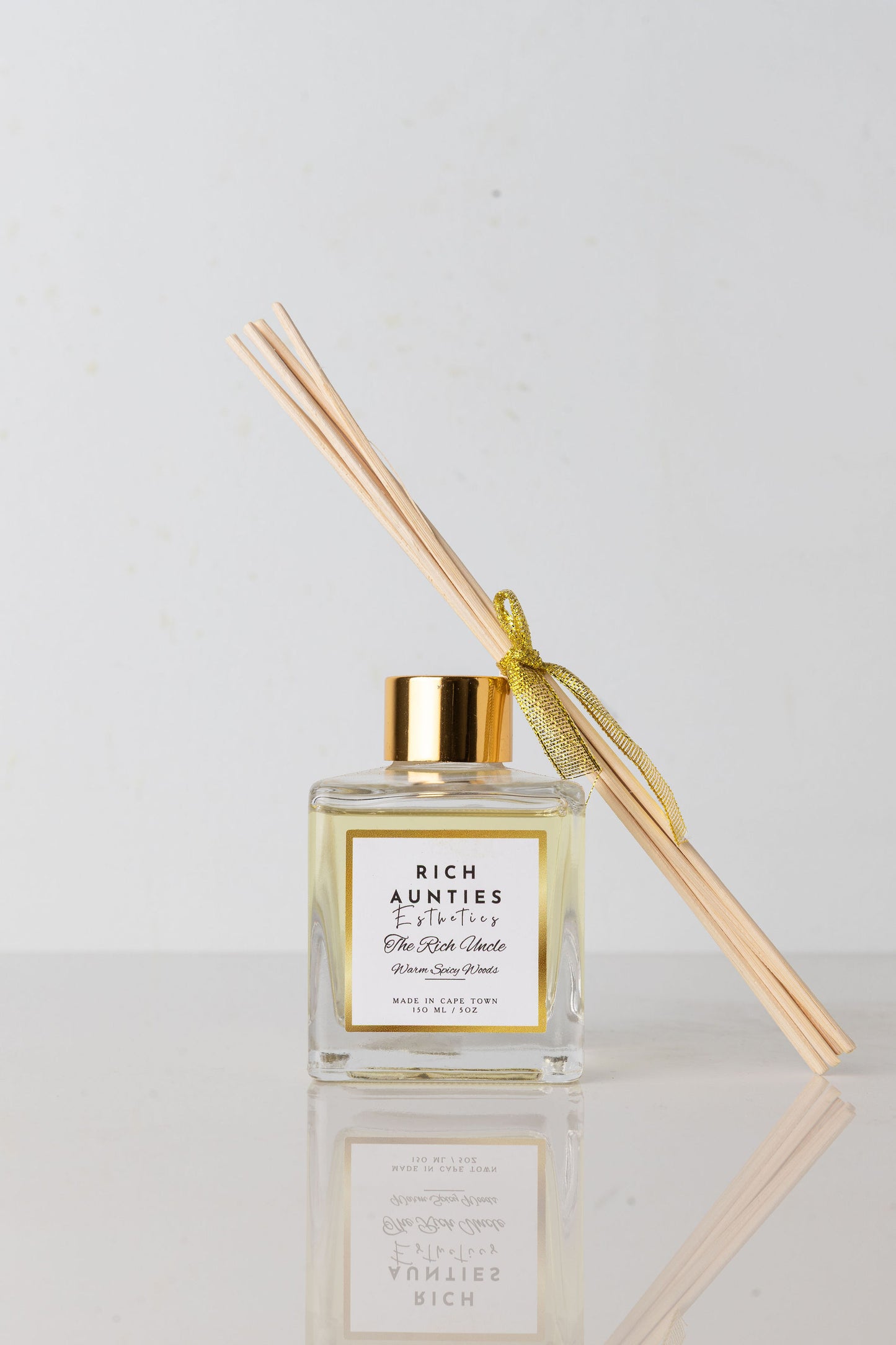 A reed diffuser with clear glass bottle and gold-colored cap, containing a diffuser fragrance and accompanied by several light beige reeds tied with a gold ribbon.