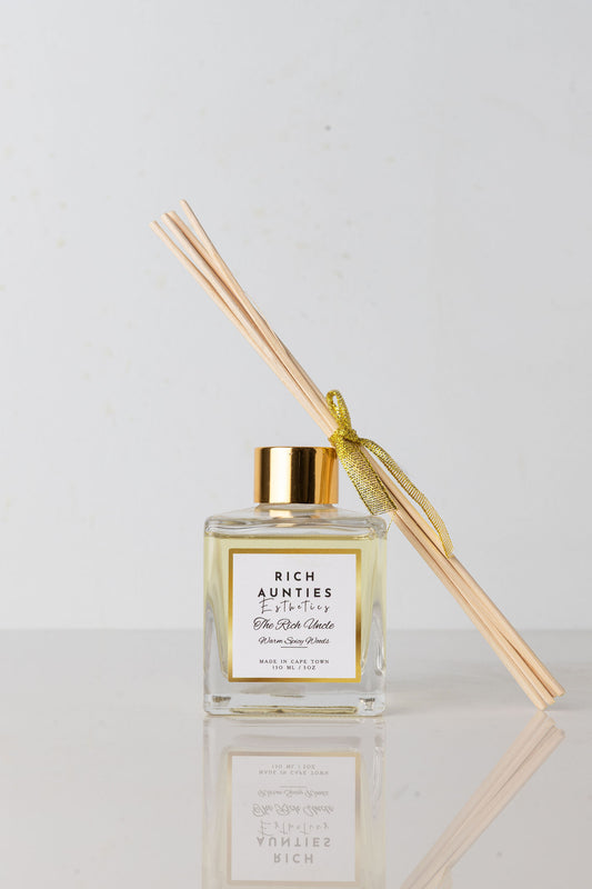 A reed diffuser with clear glass bottle and gold-colored cap, containing a diffuser fragrance and accompanied by several light beige reeds tied with a gold ribbon.