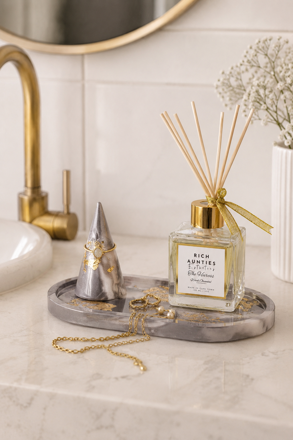Luxury reed diffuser on marble tray with gold jewelry and decorative ring holder in a modern bathroom vanity setting.