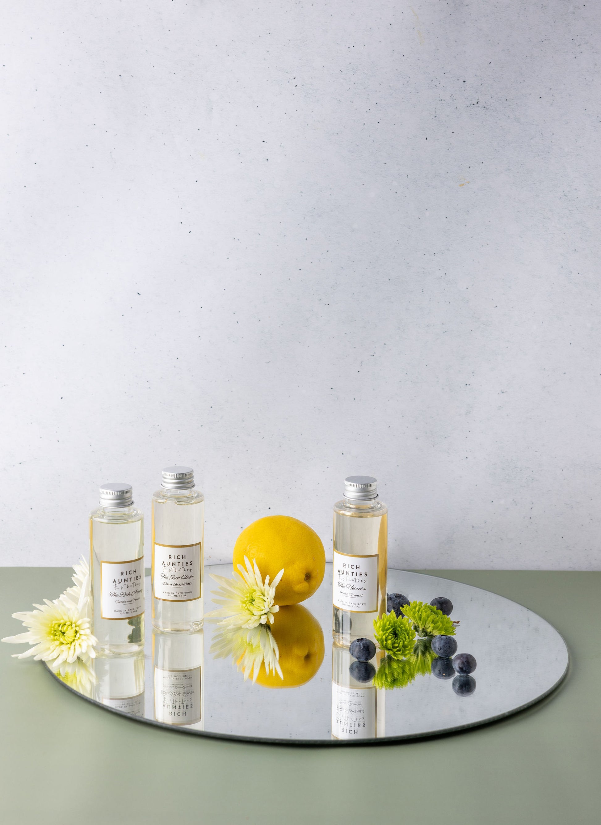Three bottles with labels on a reflective surface with a lemon and flowers on a light background